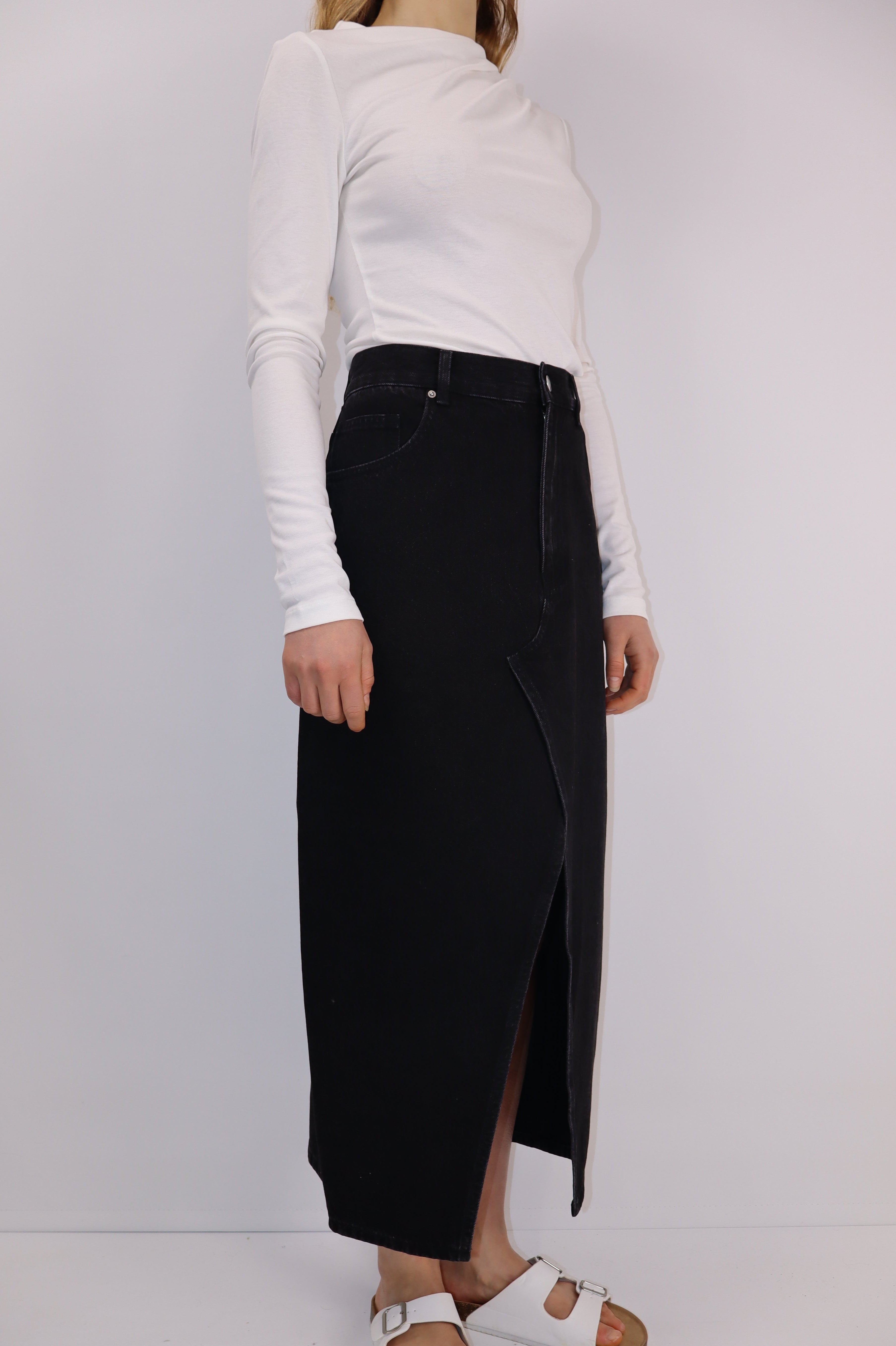 Model wearing a washed black high-waisted denim midi skirt with an asymmetrical seam and front slit, styled with a white long-sleeve top.