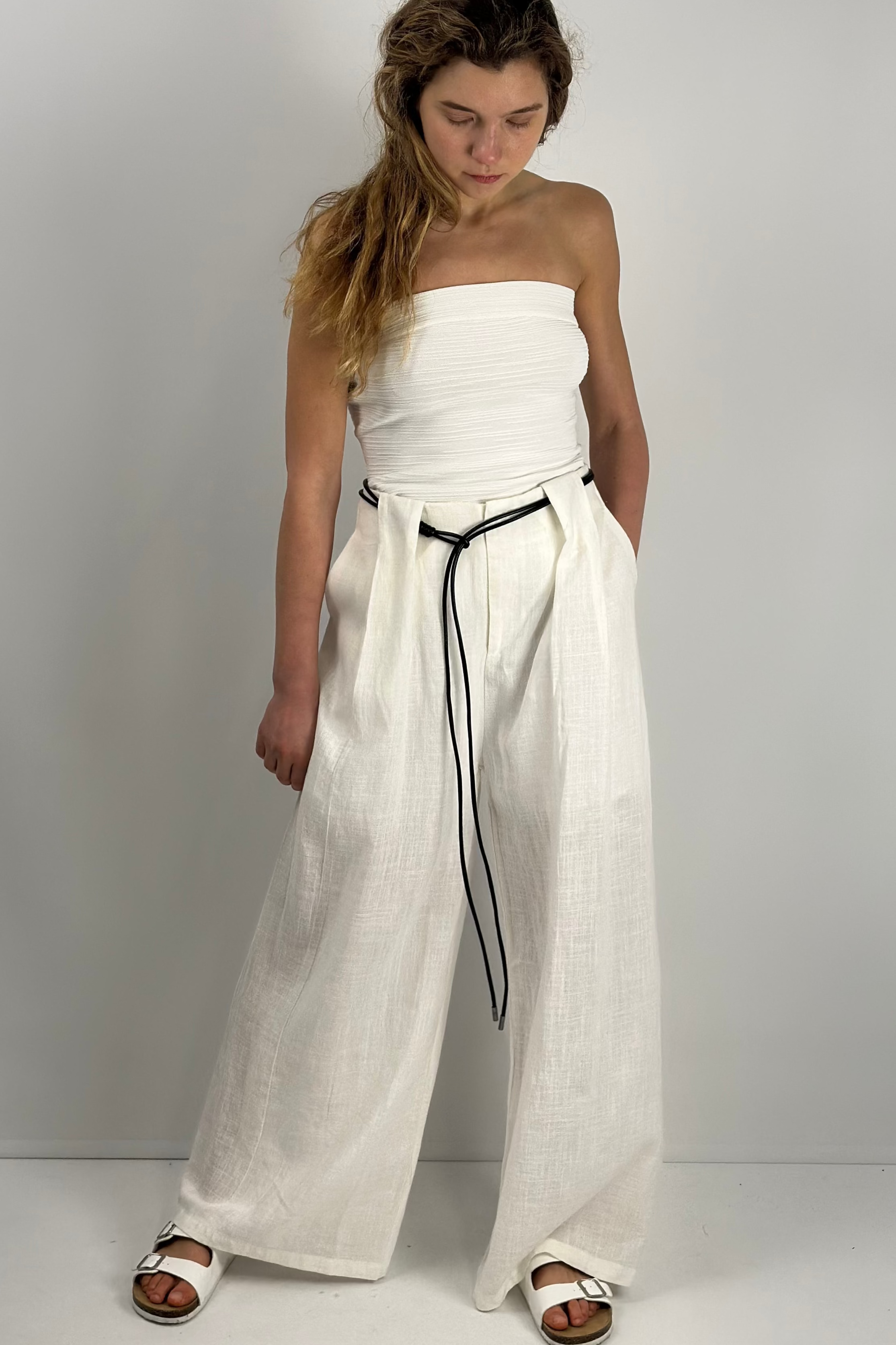 A woman wearing white wide-leg linen-Tencel blend pants with front pleats and an included slim black tie belt, styled with a black strapless top and white sandals.