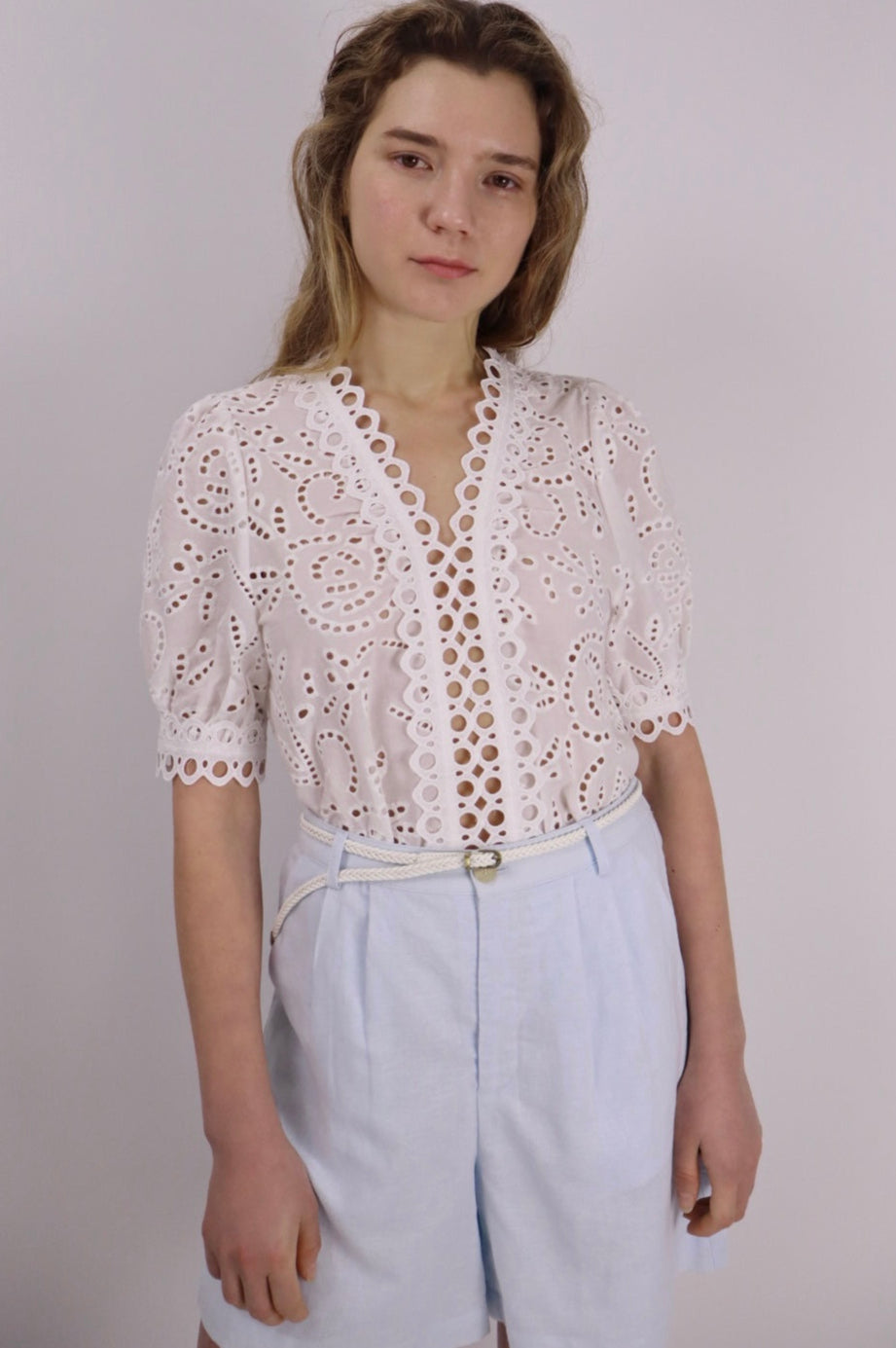 Woman wearing a white lace top and light blue pants against a plain background