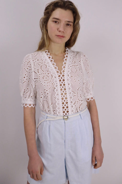Woman wearing a white lace top and light blue pants against a plain background