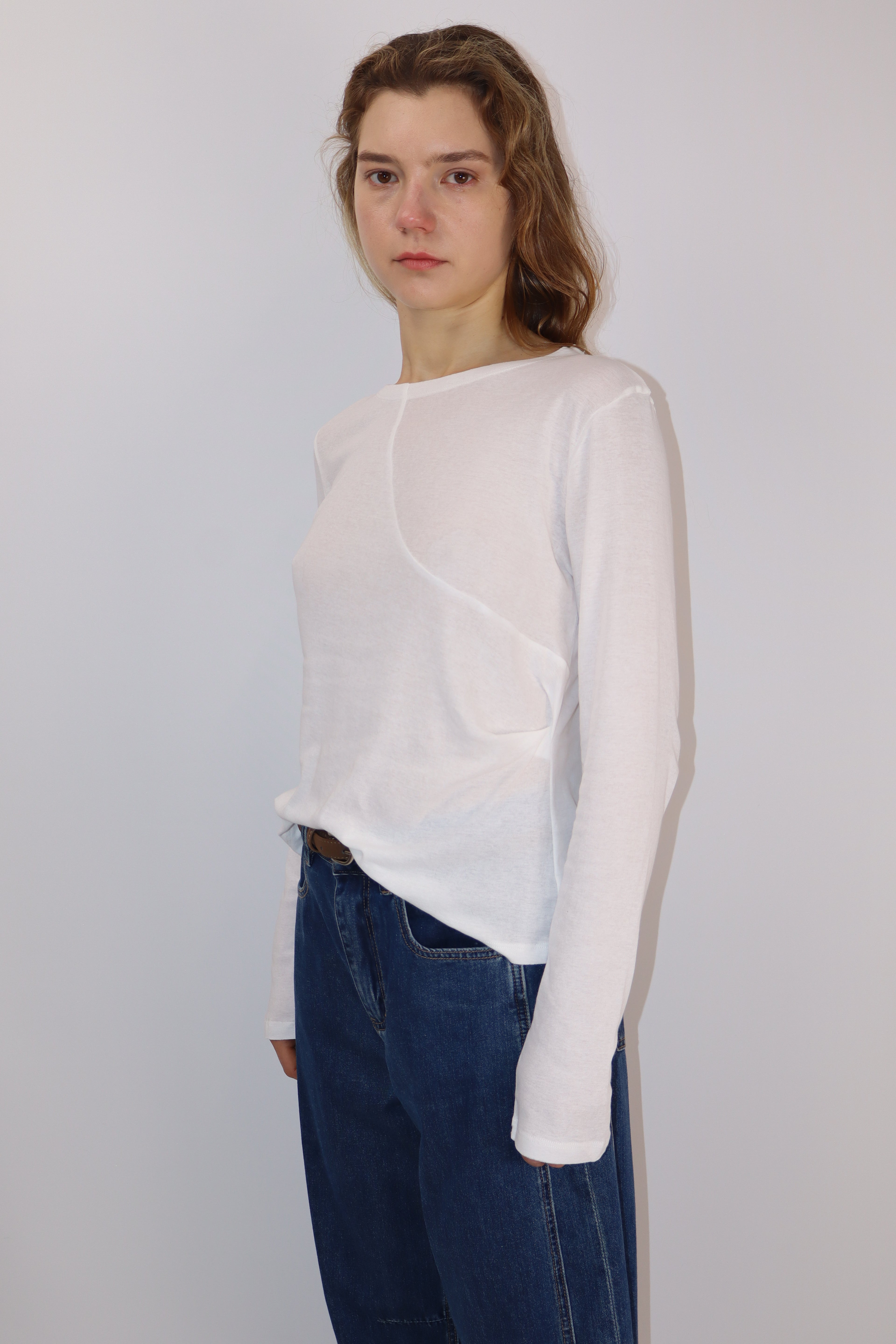 Model wearing white cotton long-sleeve top with an asymmetrical front seam and gentle gathers at the left waist, paired with blue jeans and a brown belt.