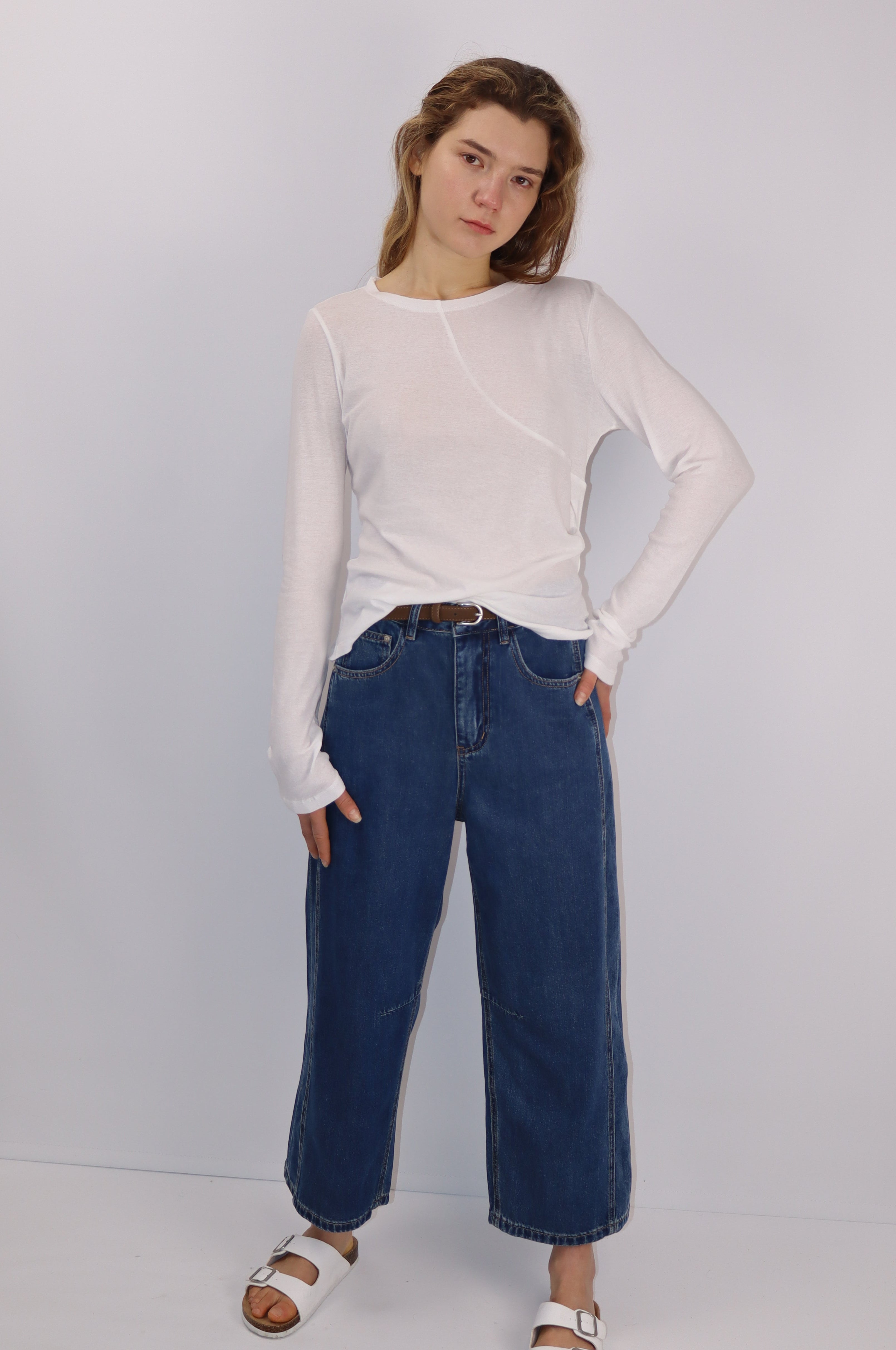 Model wearing an white cotton long-sleeve top with an asymmetrical front seam and gentle gathers at the left waist, paired with blue jeans and a brown belt.