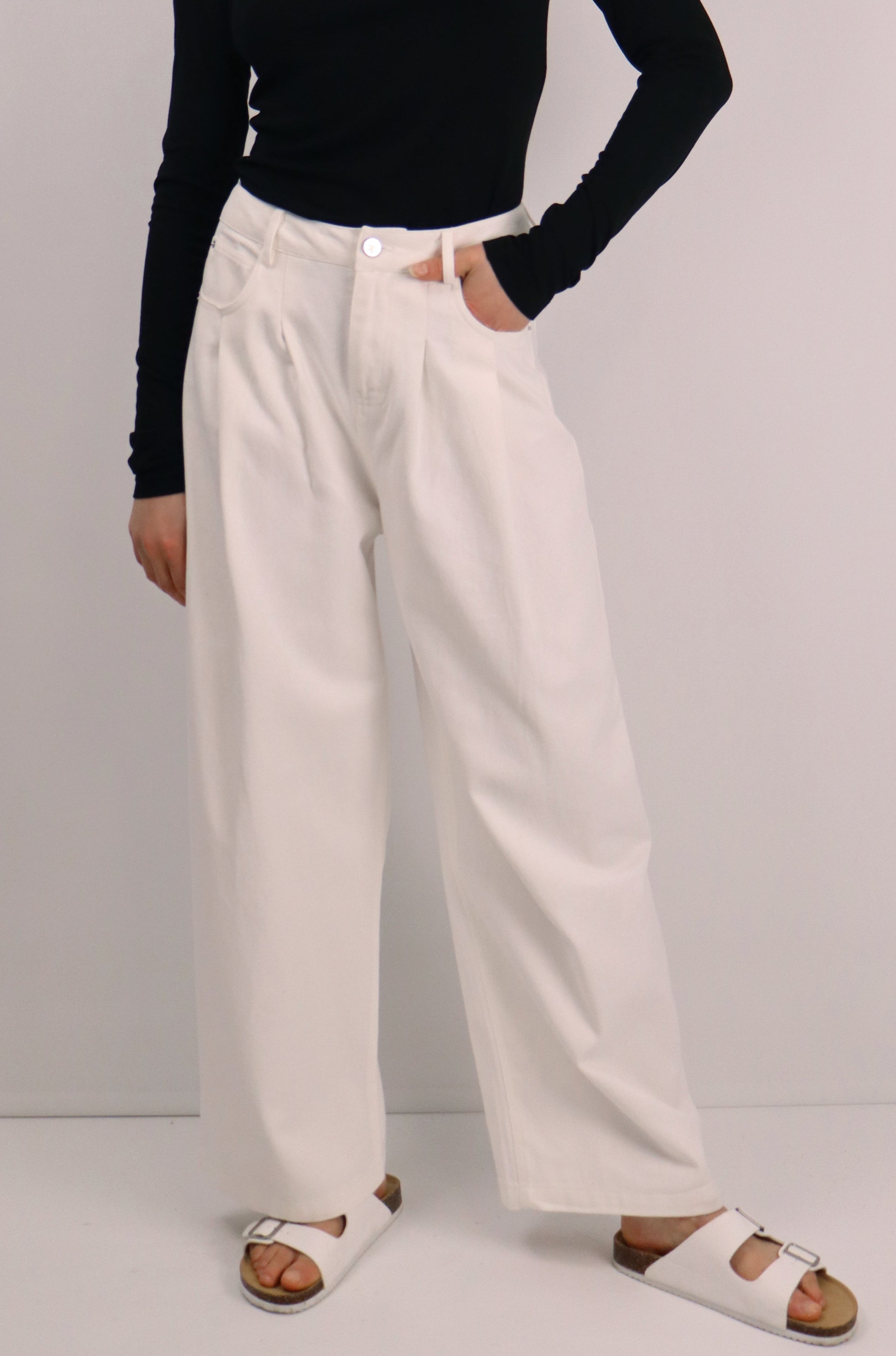 Woman wearing a black long-sleeve top and white pants against a plain background