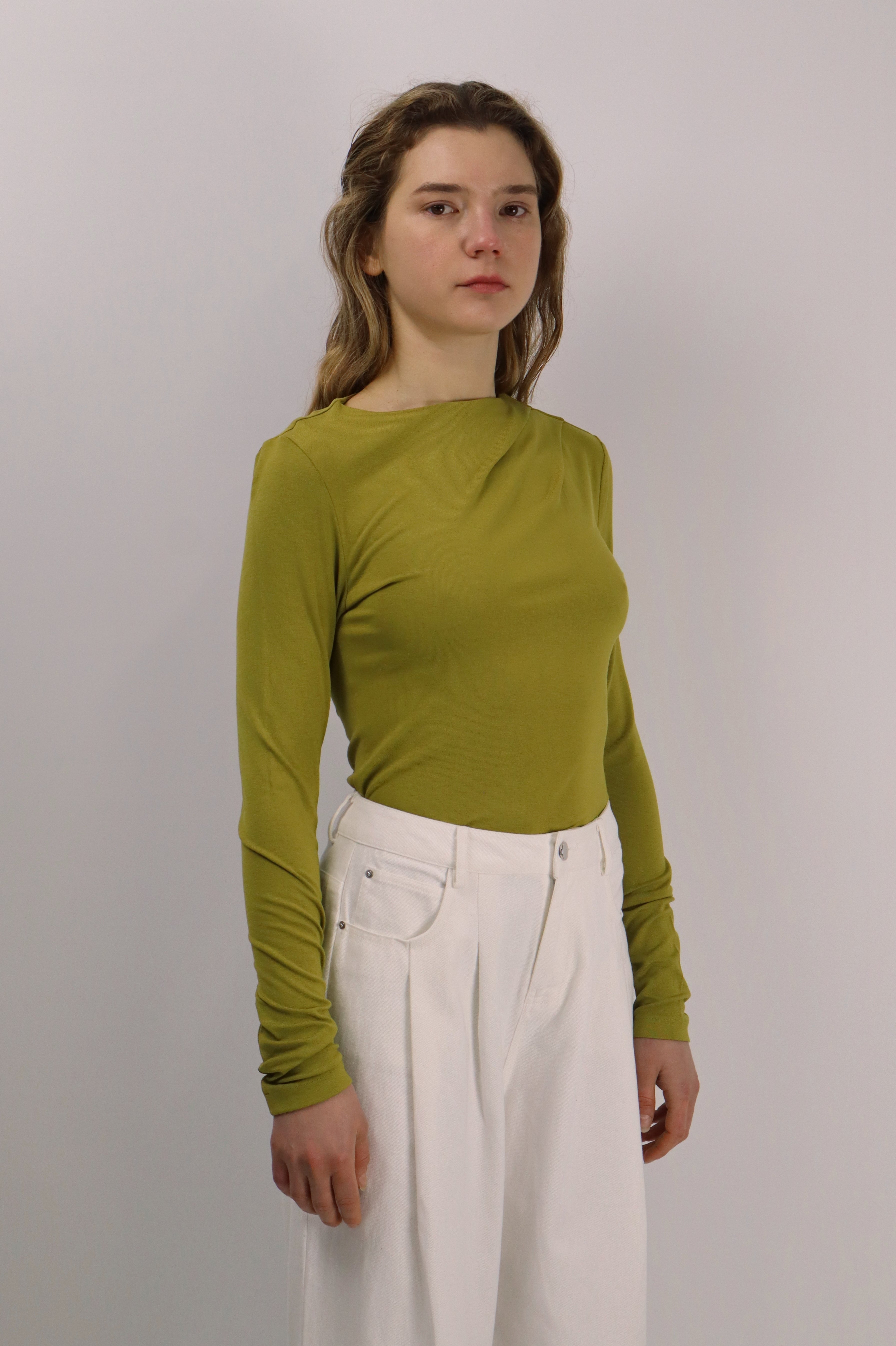 Woman wearing a green long-sleeve top and white pants against a plain background