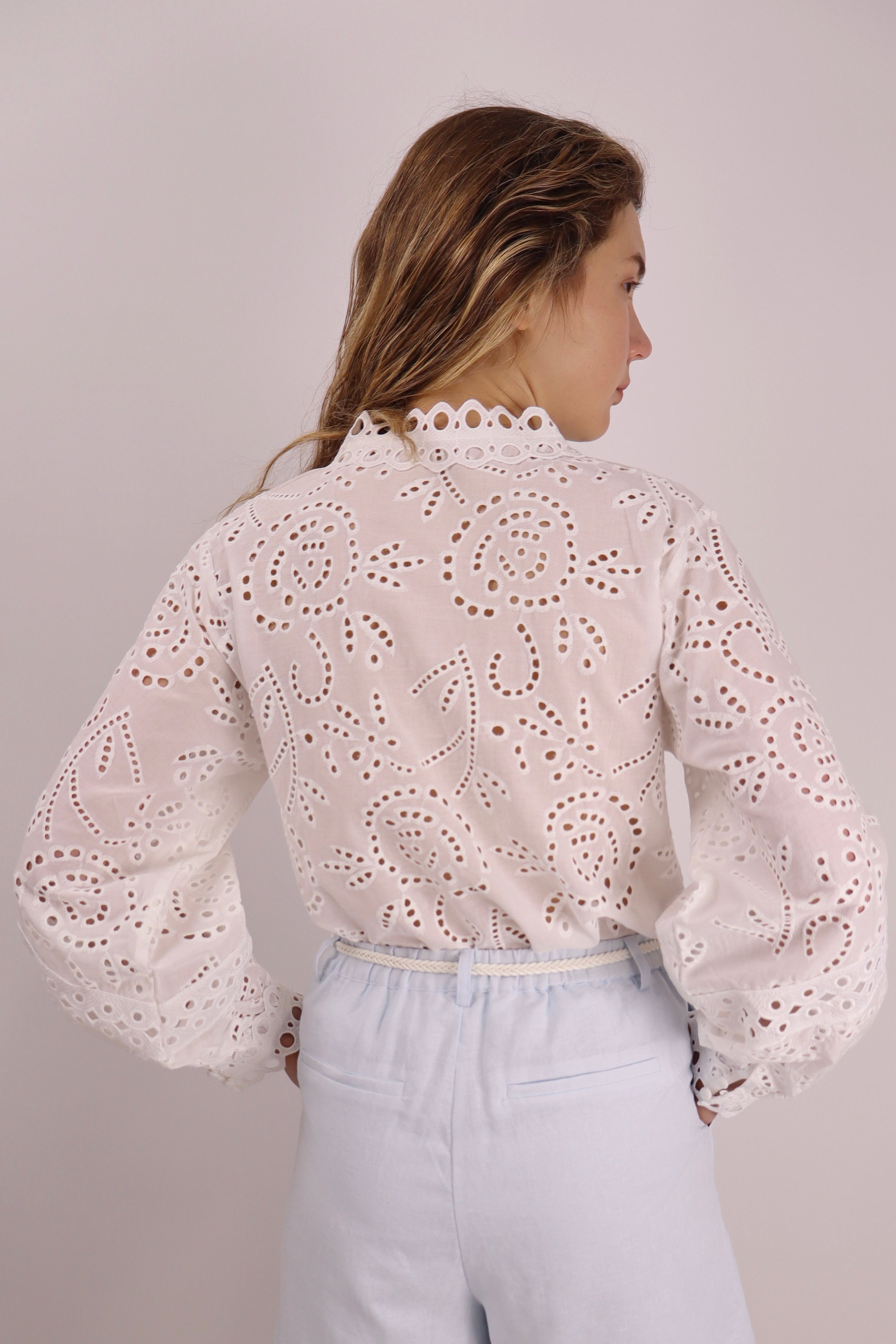Woman wearing a white lace blouse and light blue pants against a plain background