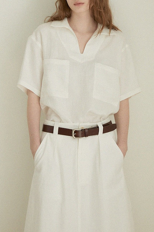 Woman wearing a relaxed white 100% linen V-neck top with short sleeves and two front pockets, styled with white trousers and a brown belt.