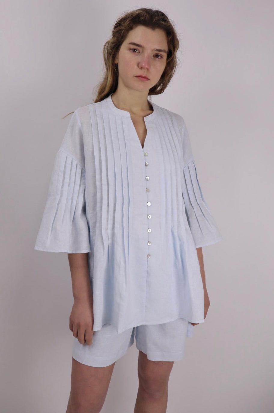 Model wearing a baby blue linen-cotton blouse with front and sleeve pintuck detailing, mandarin collar, and 3/4 wide sleeves, paired with matching white shorts, standing against a plain background.