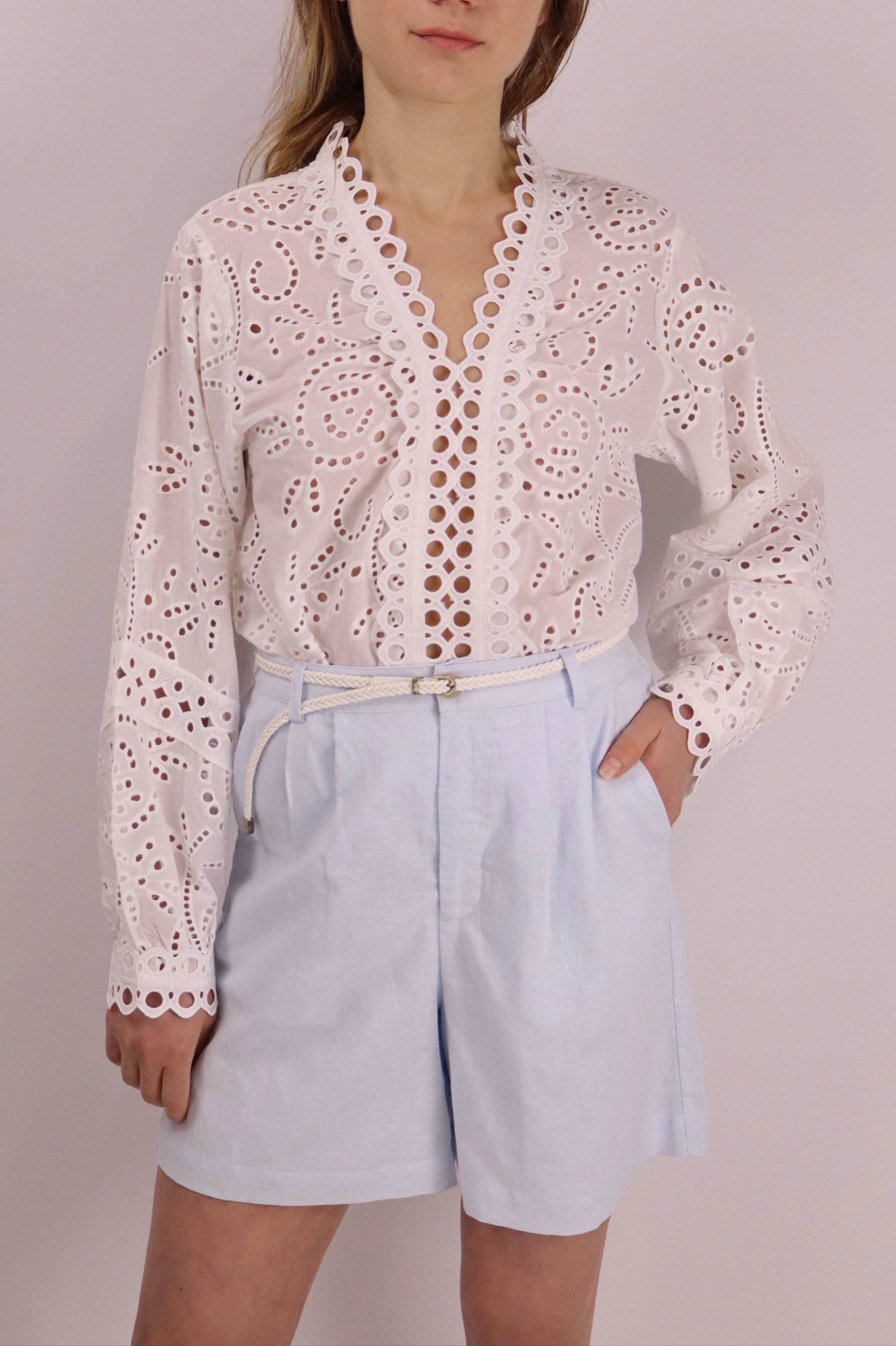 Close-up of baby blue linen-cotton shorts with front pleats, side pockets, elasticated back waistband, and a removable braided belt, worn by a model against a plain background.