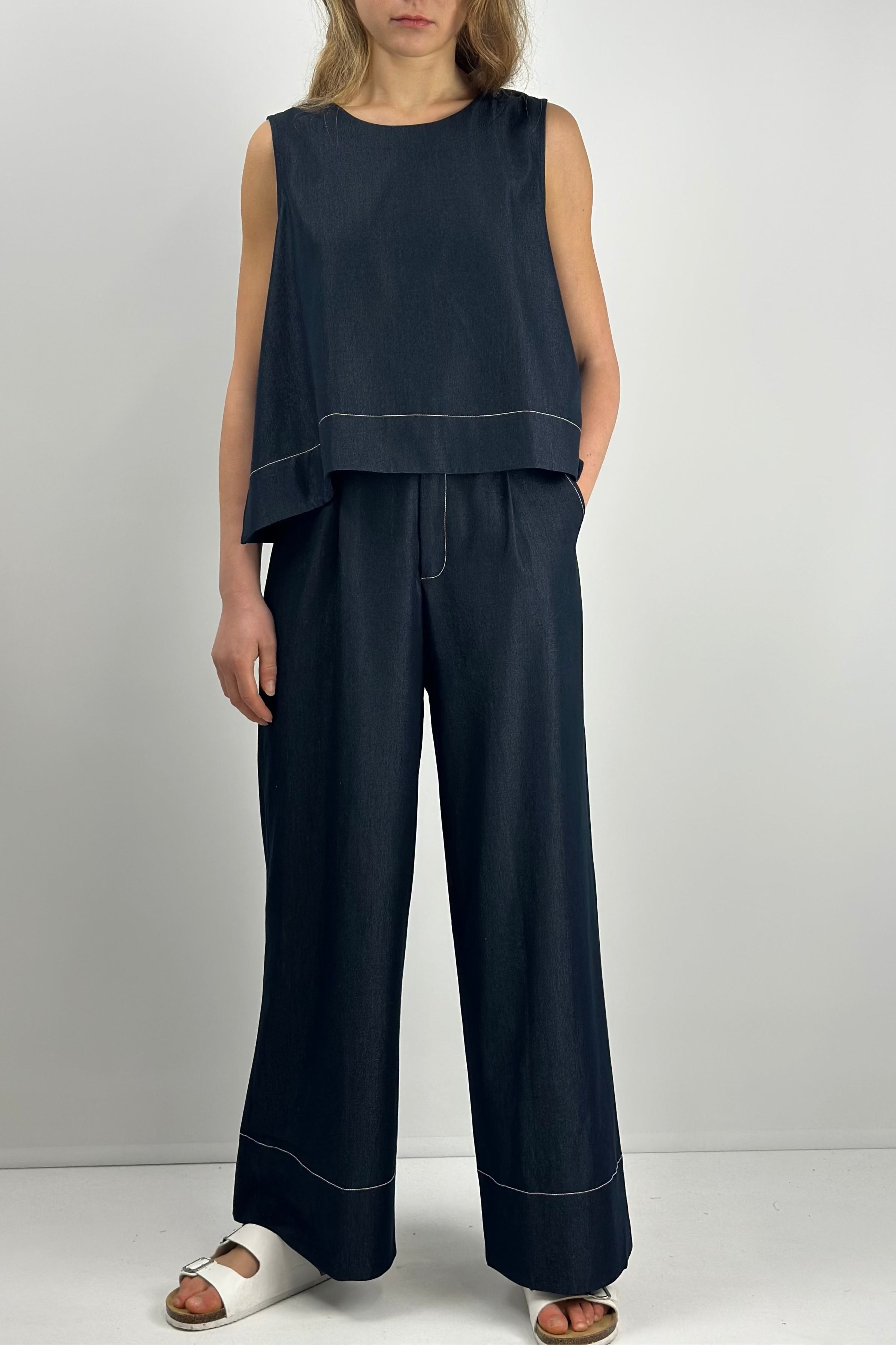 model wearing dark navy blue lightweight denim with contrast stitches wide-leg pants with matching sleeveless top and white sandals, showcasing a simple, loose, casual style.