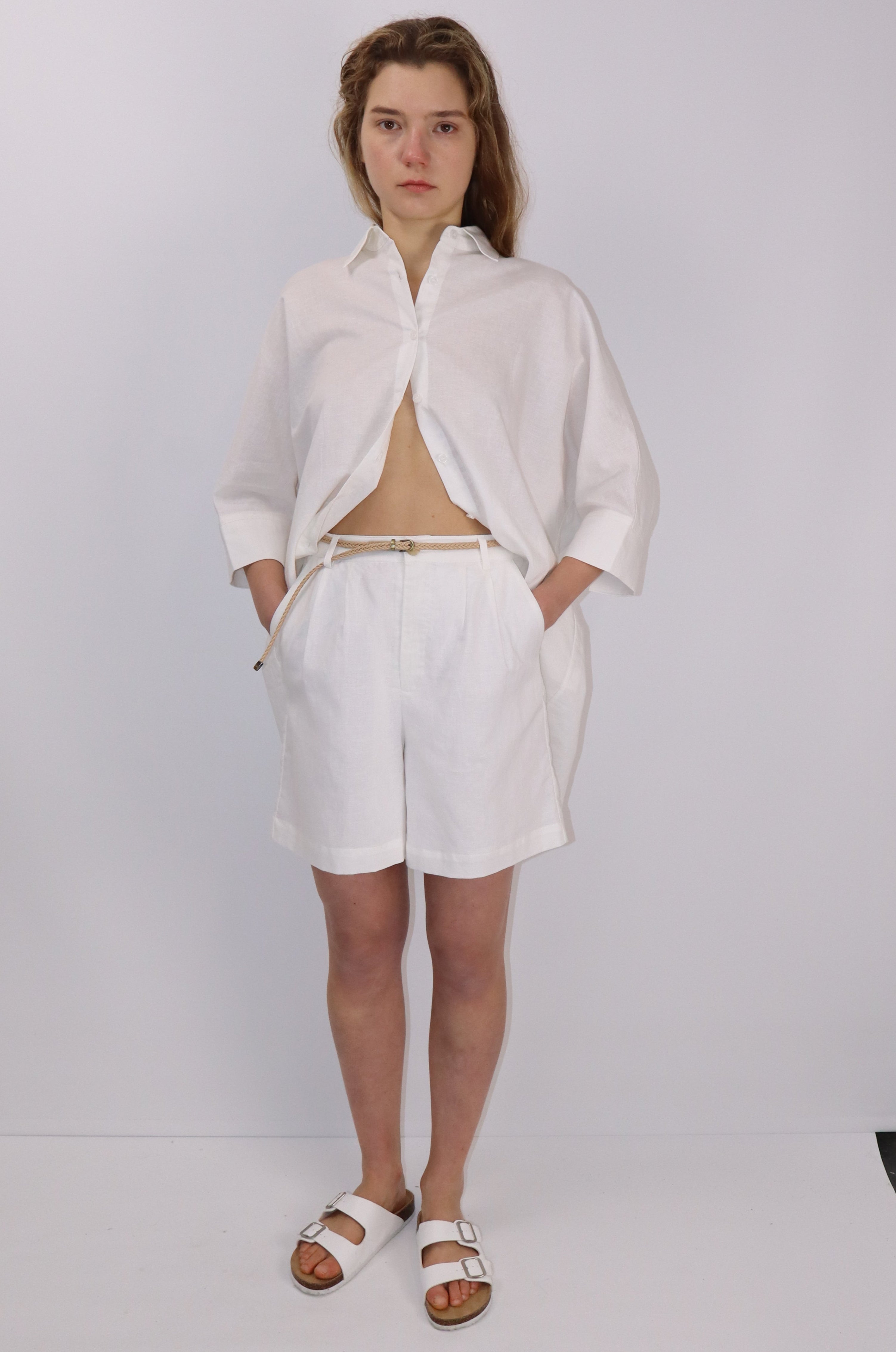 Close-up of linen-cotton shorts with front pleats, side pockets, elasticated back waistband, and a removable braided belt, worn by a model against a plain background.