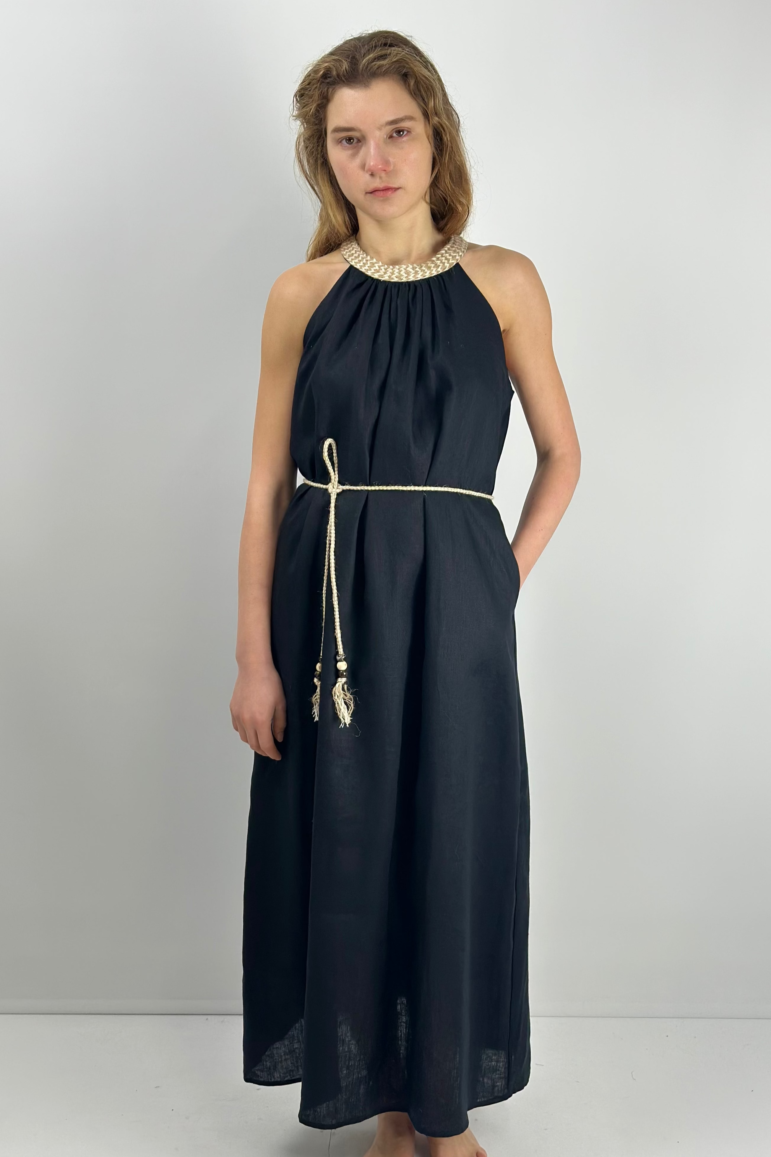 Model wearing a black linen maxi dress with braided neckline and matching braided belt, hands in pockets, barefoot.