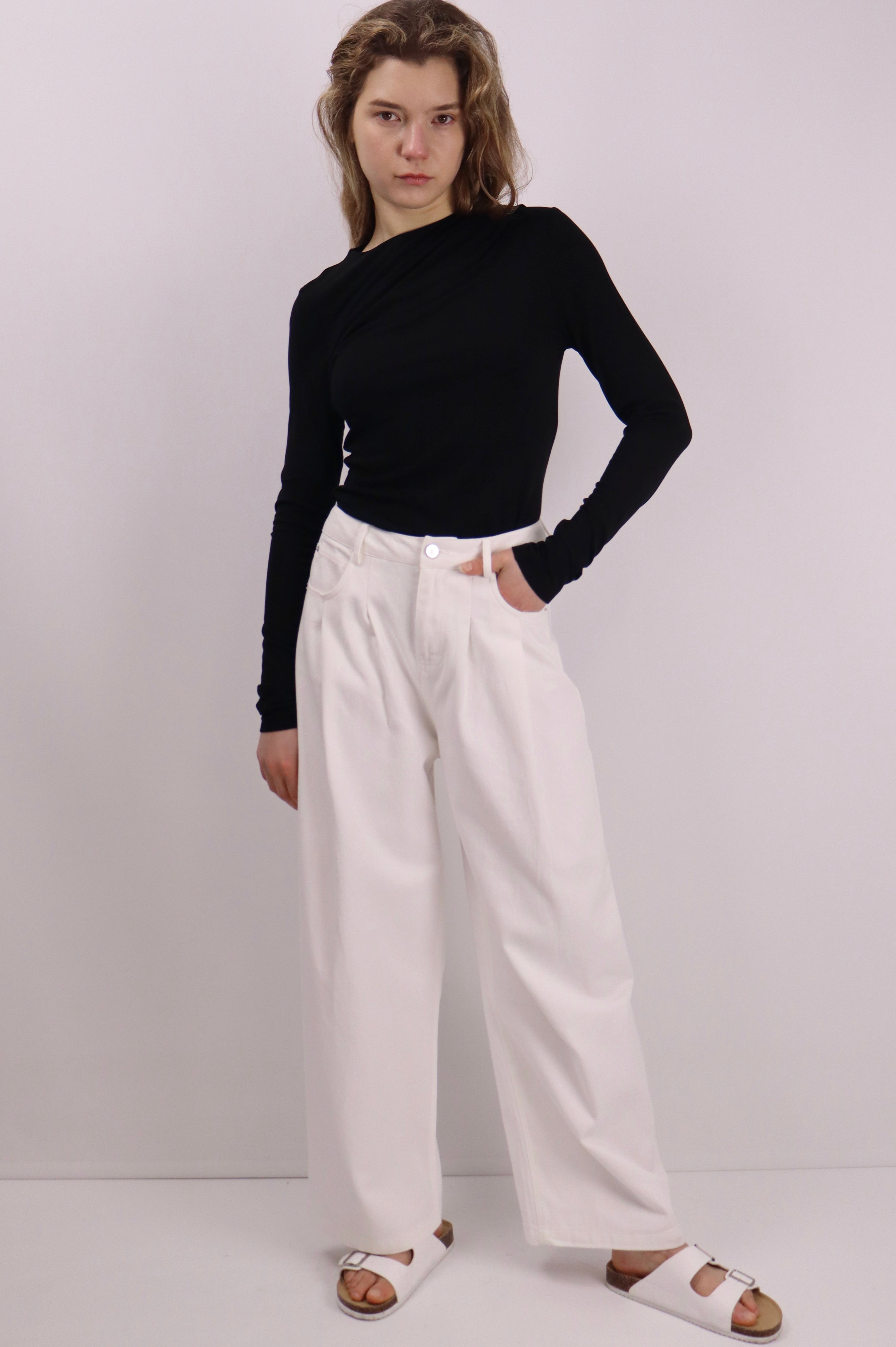 Model wearing mid-rise wide-leg white cotton pants with front pleats, paired with a fitted black long-sleeve top and white sandals, standing against a plain background.