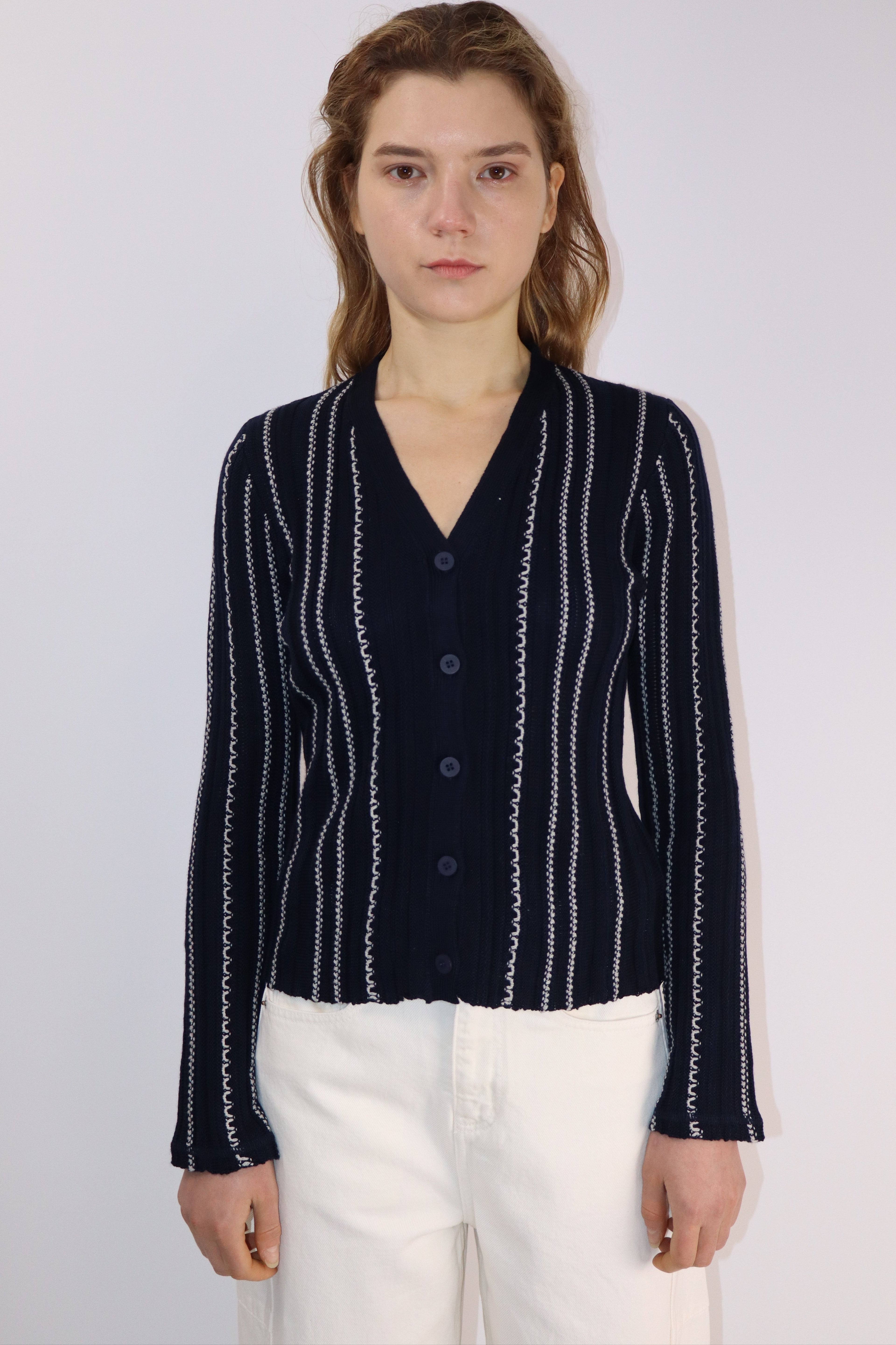 Model wearing a fitted navy cardigan with vertical white stripes and button-up front, paired with off-white wide-leg pants, standing against a plain light background.