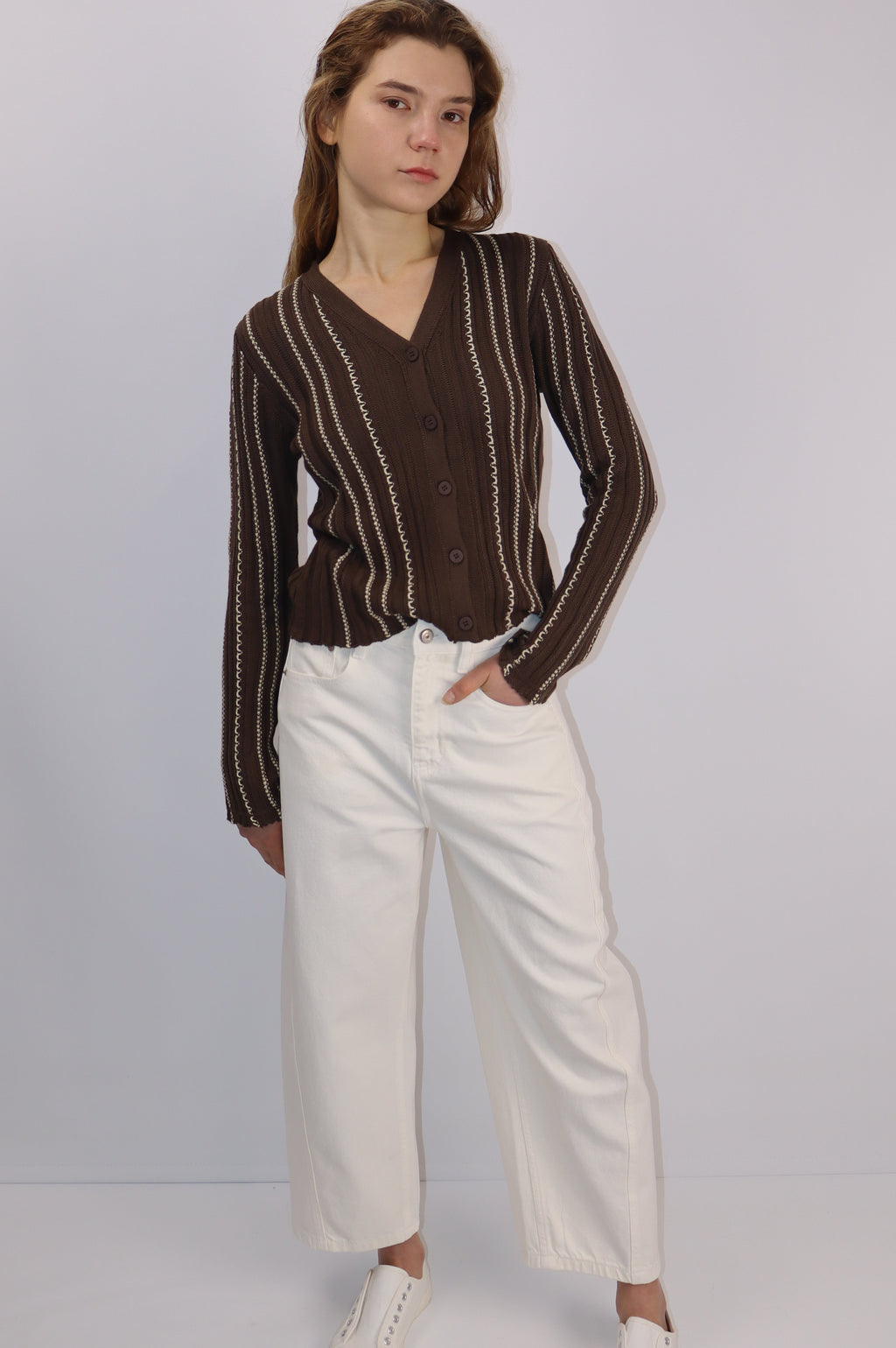 Woman wearing a brown striped cardigan and white pants on a plain background