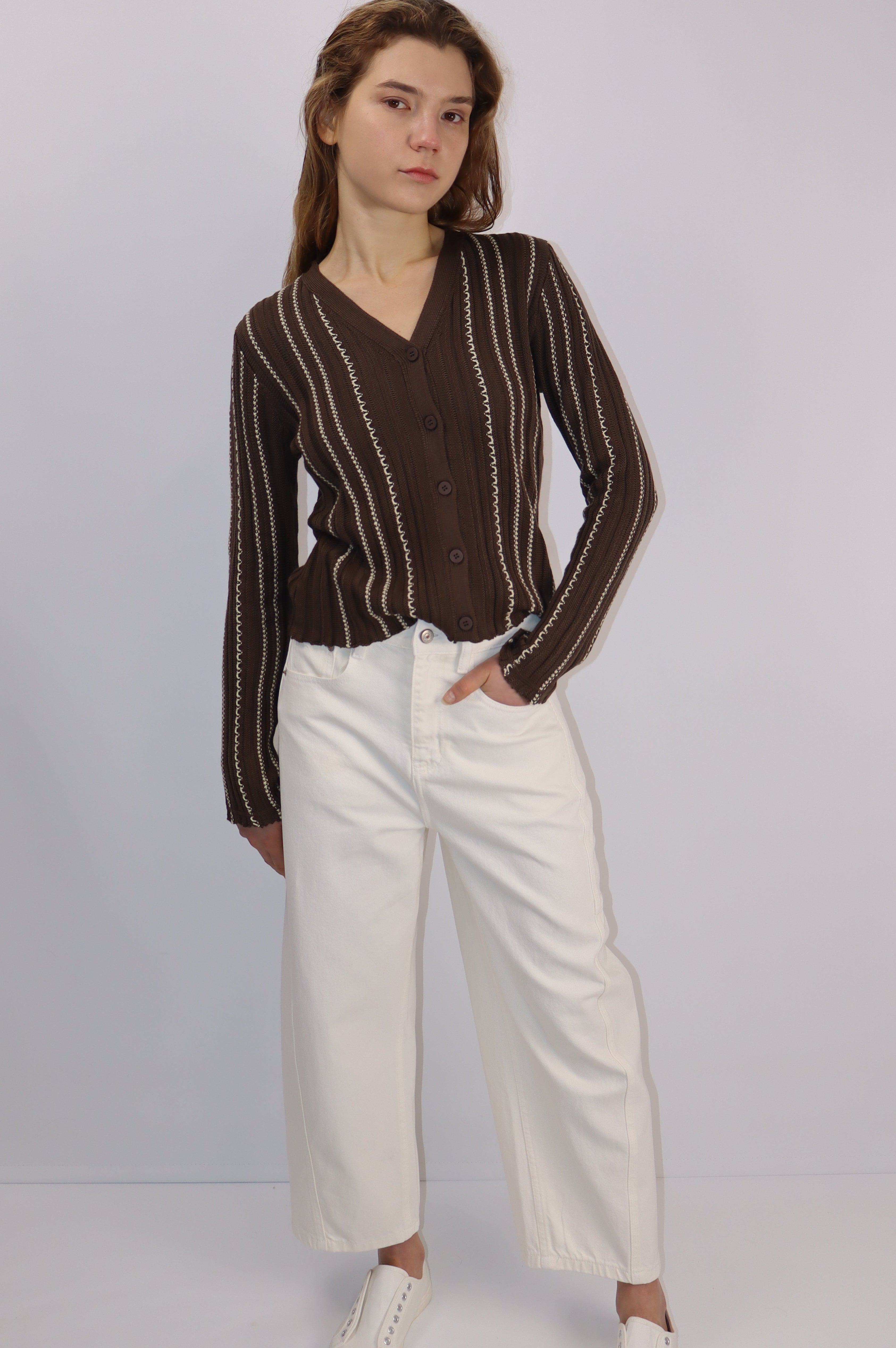 Woman wearing a brown striped cardigan and white pants on a plain background