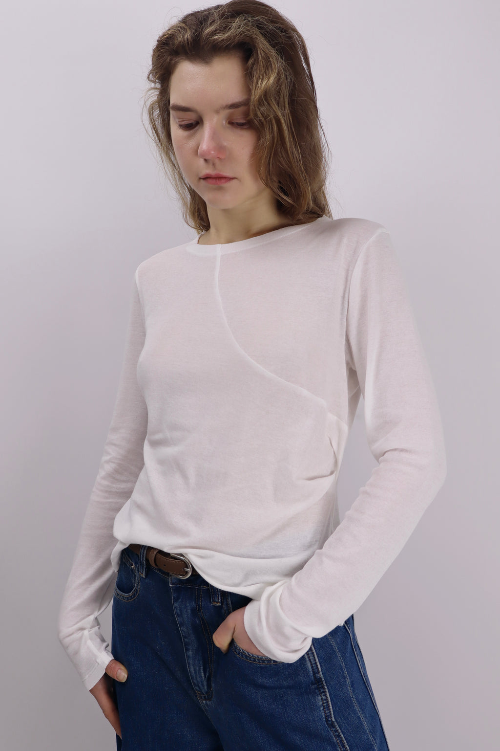 Model wearing an off-white cotton long-sleeve top with an asymmetrical front seam and gentle gathers at the left waist, paired with blue jeans and a brown belt.