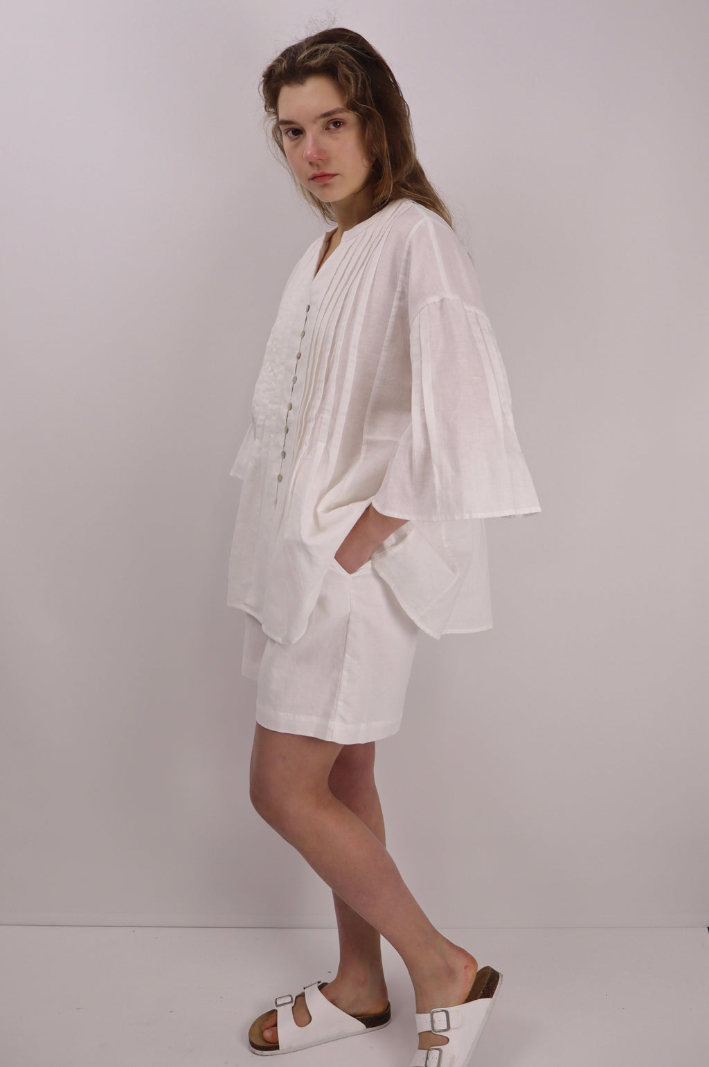 Woman wearing a white linen blouse with pleats details and white sandels against a plain background