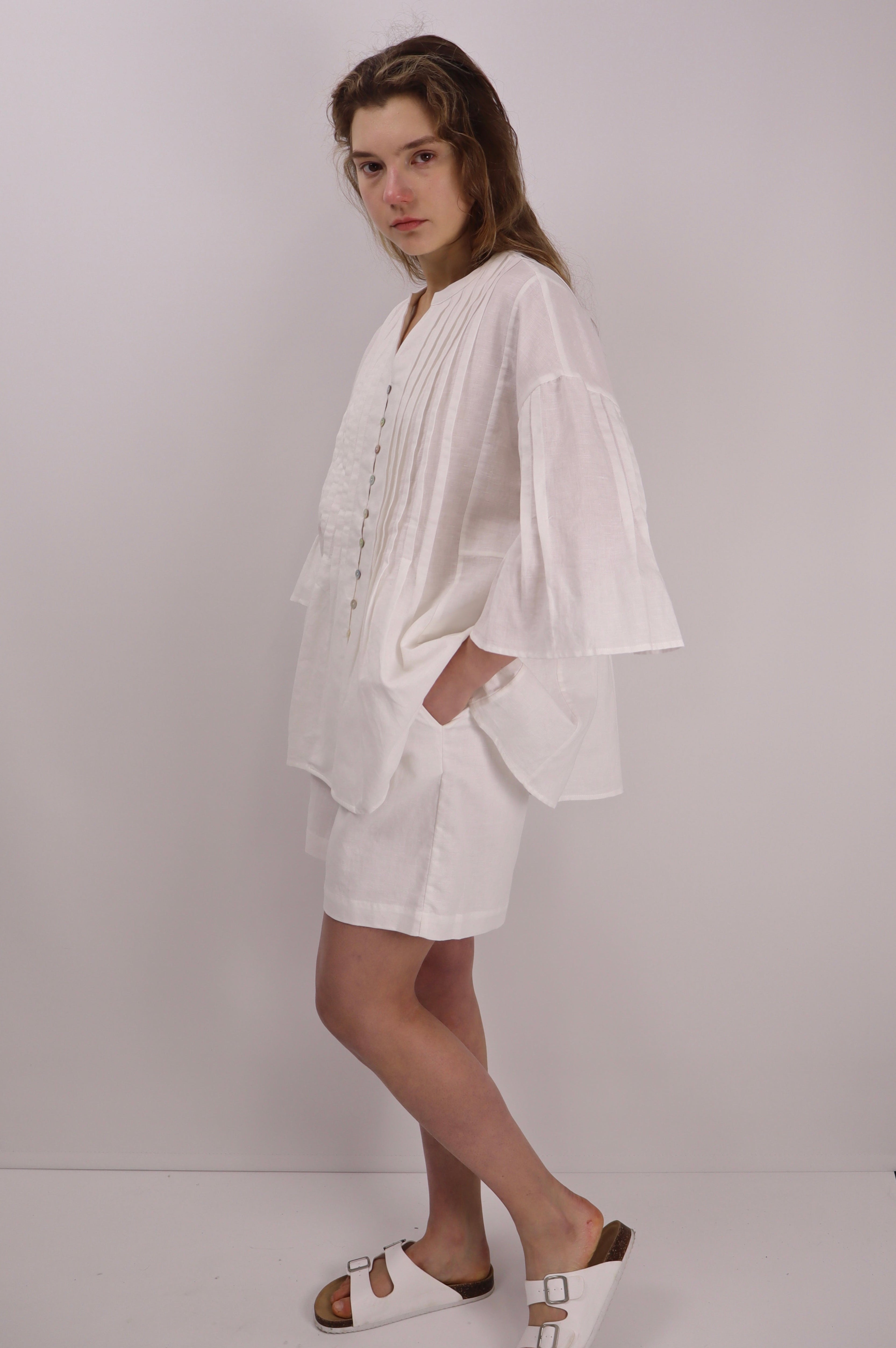 Woman wearing a white linen blouse with pleats details and white sandels against a plain background