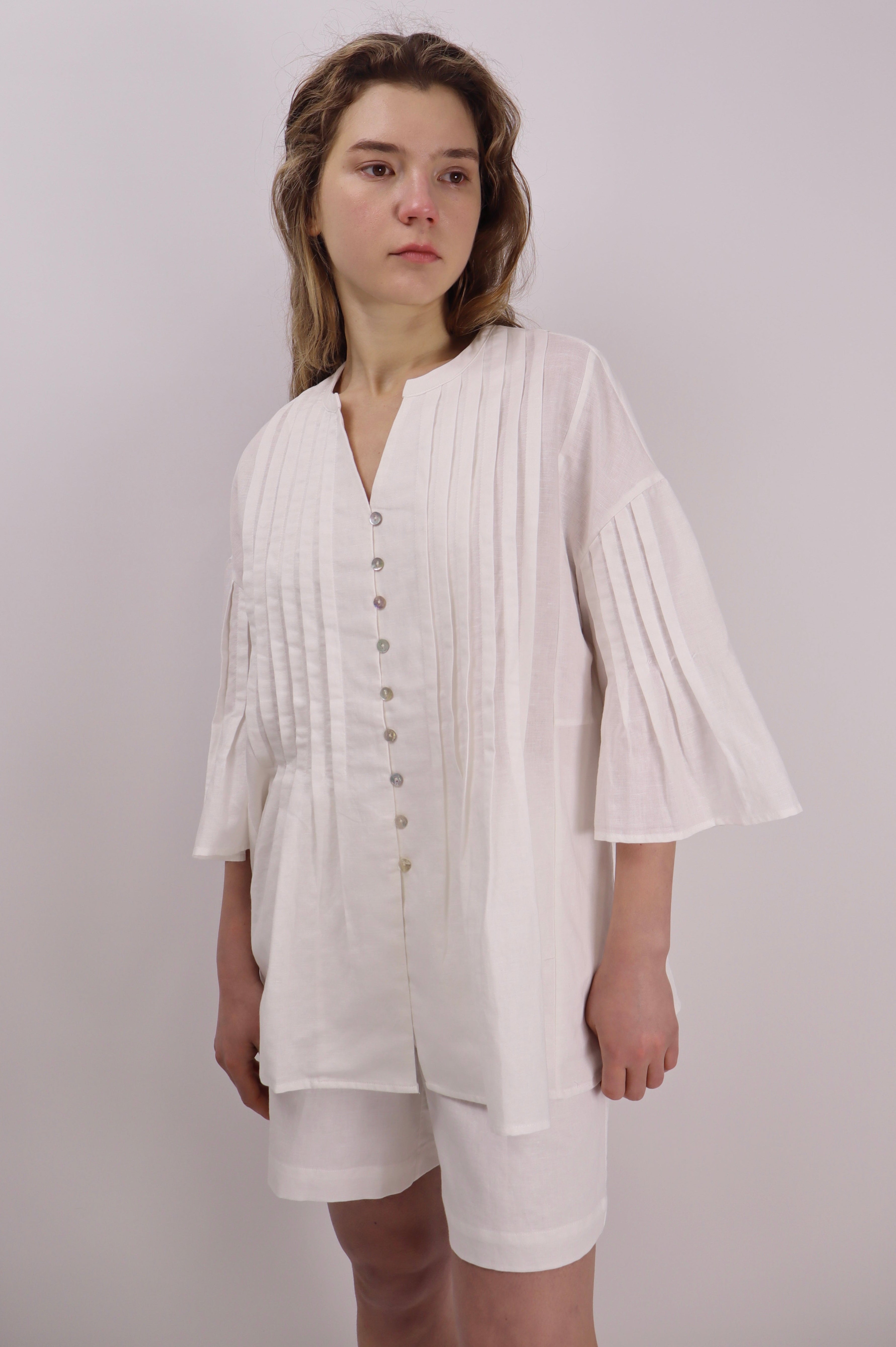 Woman wearing a white linen blouse with pleats and buttons front with a plain background