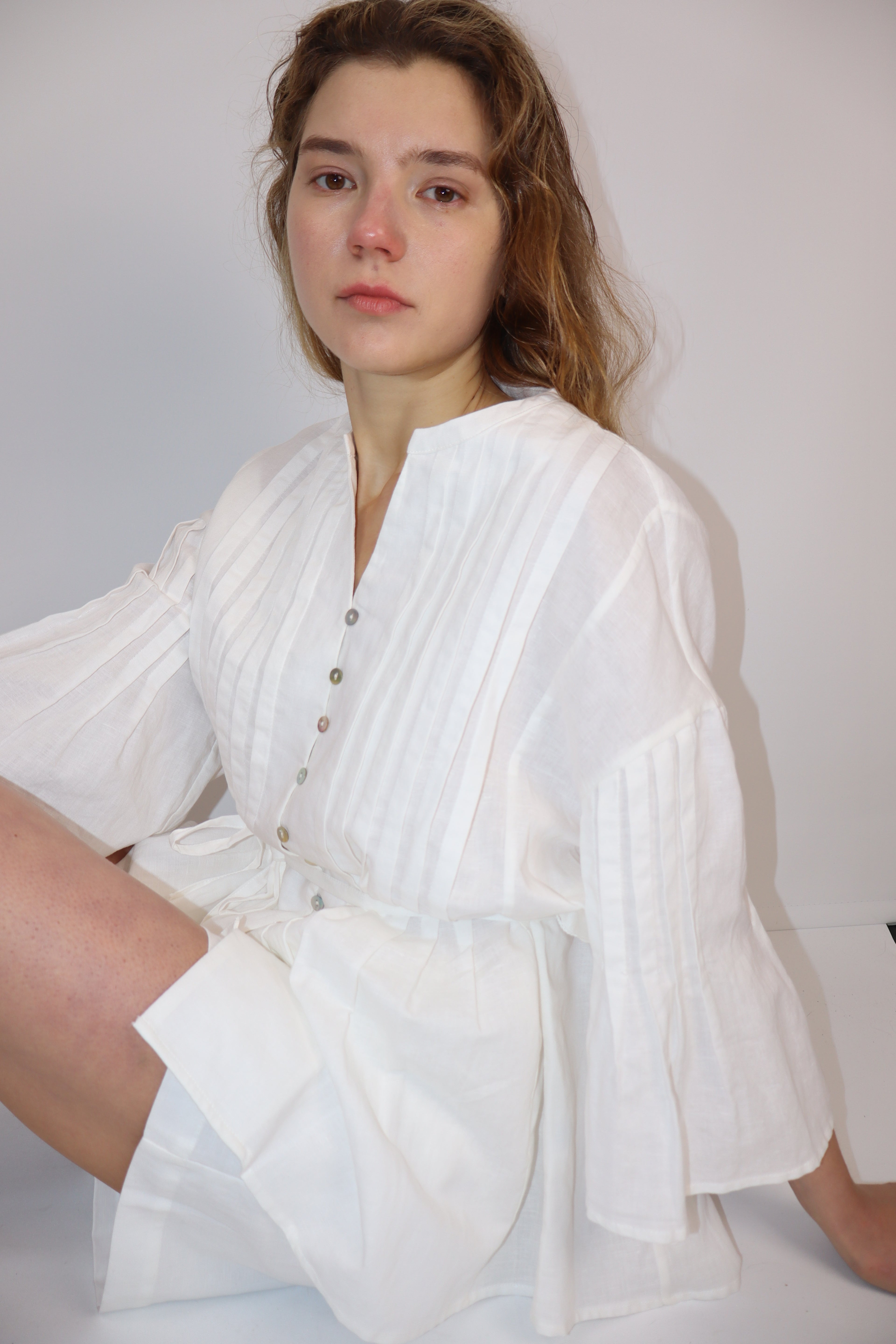 Woman sits down wearing a white linen button front blouse with tie and  pleats and against a plain background