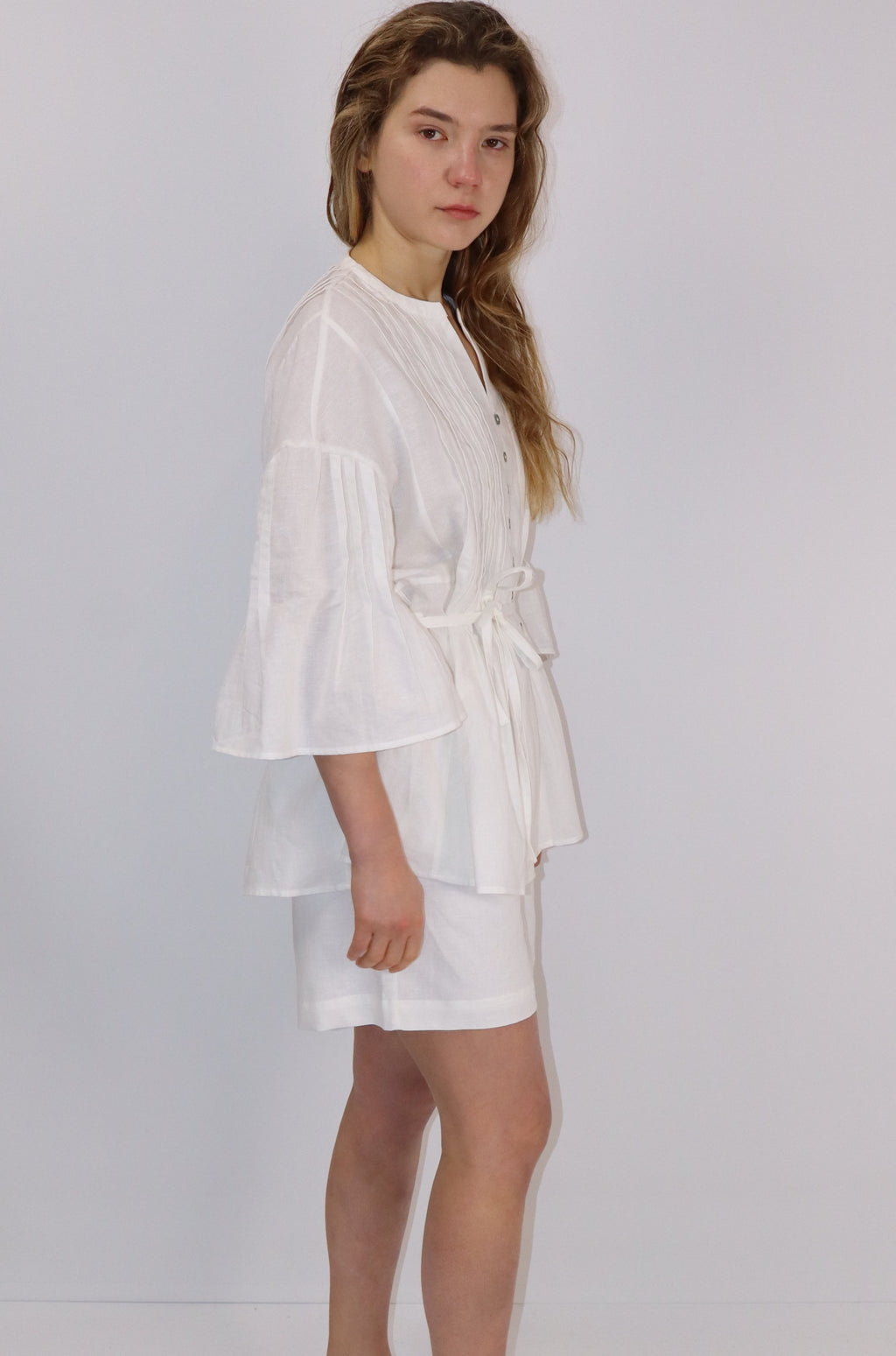 Woman wearing a white linen button front blouse with tie and  pleats and against a plain background